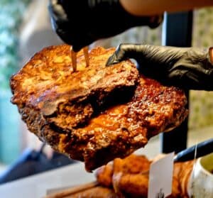 יד עטויה בכפפה אוחזת במזלג חתיכה גדולה ועסיסית של בשר מבושל מעל שולחן עמוס אוכל.